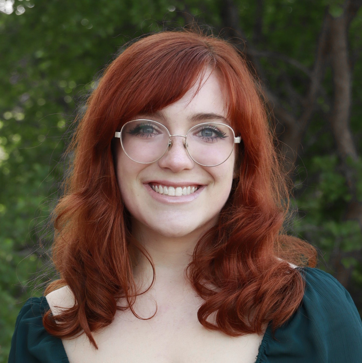 Professional portrait of H-STAR leader and current Michigan State University student Katherine Dyal smiling in a dark green top. Lush greenery is in the background.