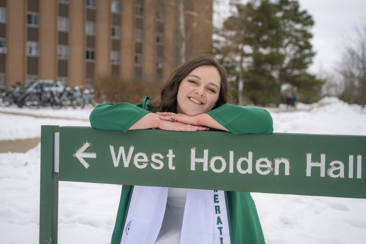Image of Michigan State University Honors College student Cheyenne Marchand in a green graduation gown, smiling and leaning on a long green sign that days "West Holden Hall". Behind Marchand is a large brick building, pine trees, and snowy ground.