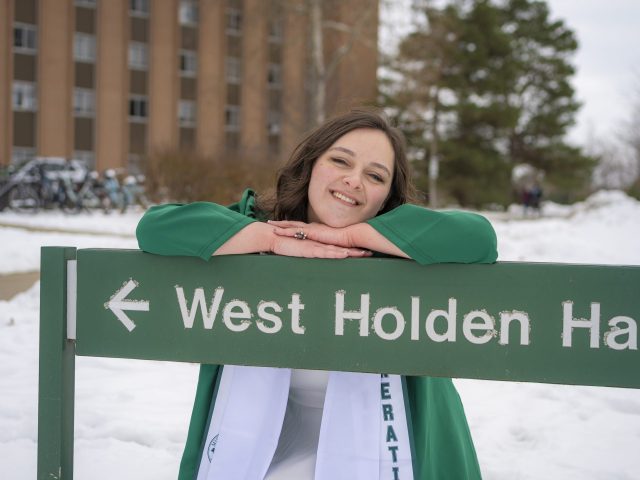 Image of Michigan State University Honors College student Cheyenne Marchand in a green graduation gown, smiling and leaning on a long green sign that days "West Holden Hall". Behind Marchand is a large brick building, pine trees, and snowy ground.