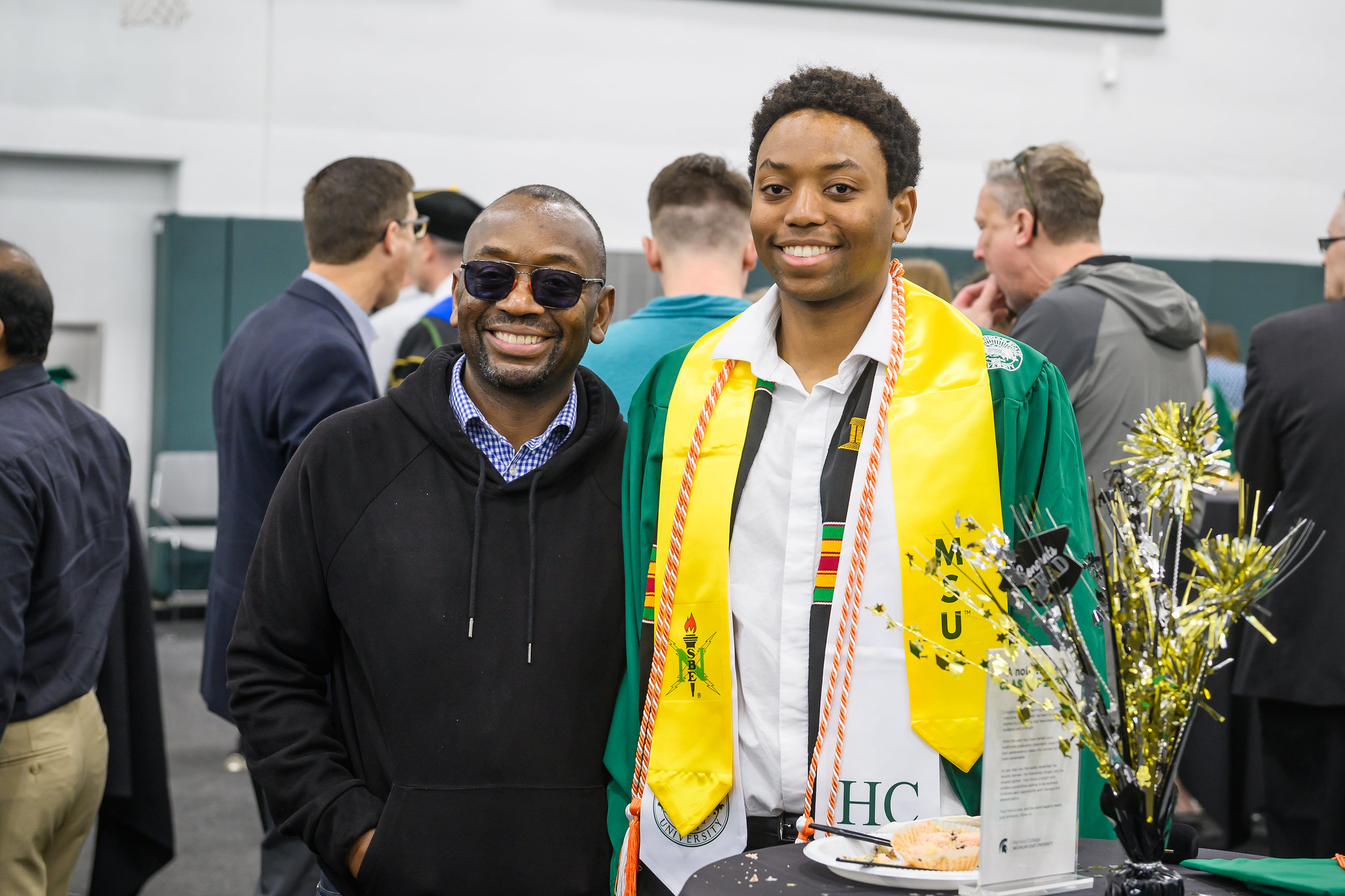 Image of a Michigan State University Honors College graduate and supporter standing and smiling at the Honors College Graduation Reception in the Breslin Center. The graduate is in a dark green gown, white collar stole, and yellow collar stole. The supporter is wearing a black hooded sweatshirt with a blue collared shirt underneath.