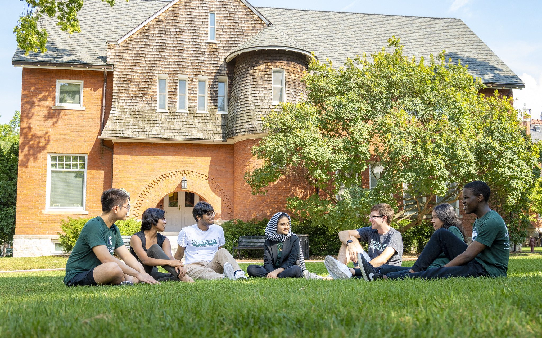 Image of six honors students in msu and honors clothing sitting on the lawn in front of Eustace Cole hall on a sunny day, smiling and laughing.