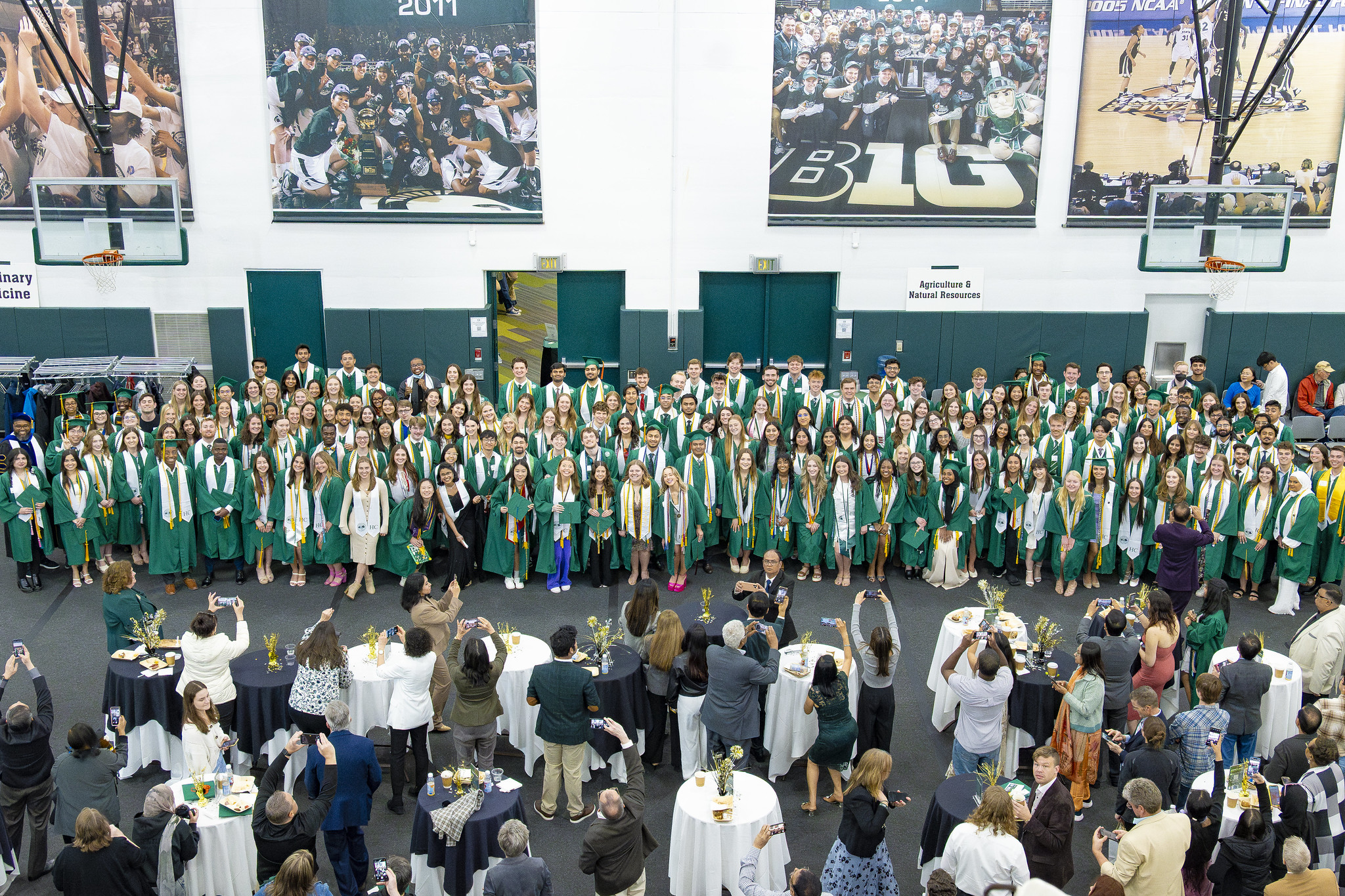Aerial view group photo of Michigan State University Honors College spring graduates in the Breslin Center. They are smiling and wearing green graduation gowns and collar stoles. A group of supporters is standing in front of them, taking photos of the graduates.