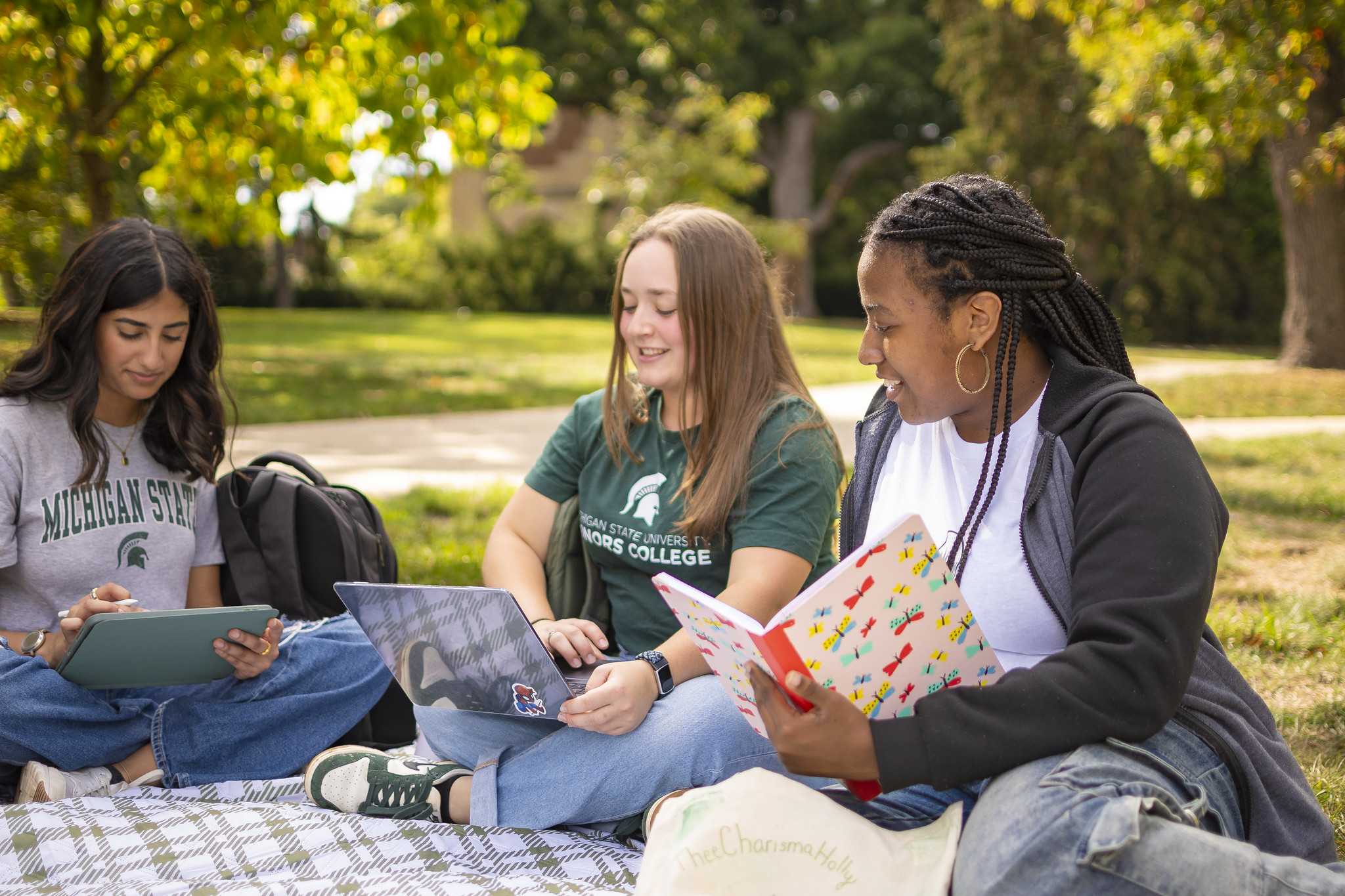 Image of three Honors students sitting on a picnic blanket working on laptops and notebooks outdoors and smiling.