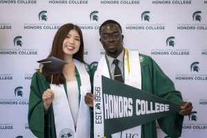 Image of two Honors graduates in dark green graduation gown and white collar stoles. They are holding photo booth props smiling in front of a white background with a repeating patter of green Michigan State University Honors College logos.