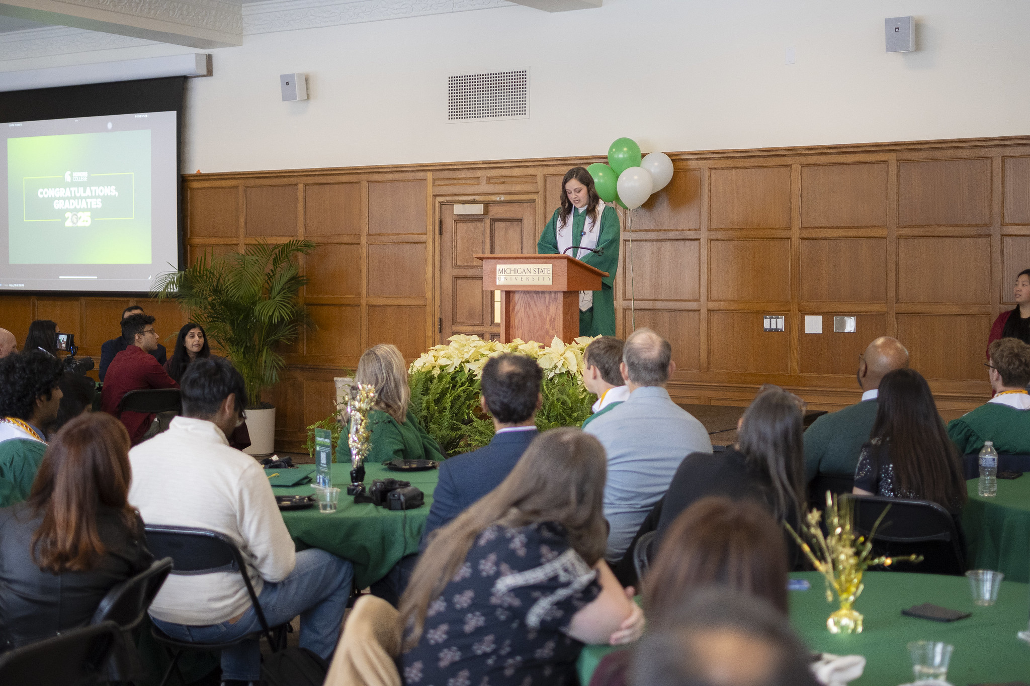 Image of a Michigan State University Honors College graduate delivering speech in Campbell Hall to a crowd of people. The graduate is in a green gown and white collar stole. A wood paneled wall and small bunch of green and white balloons are in the background.