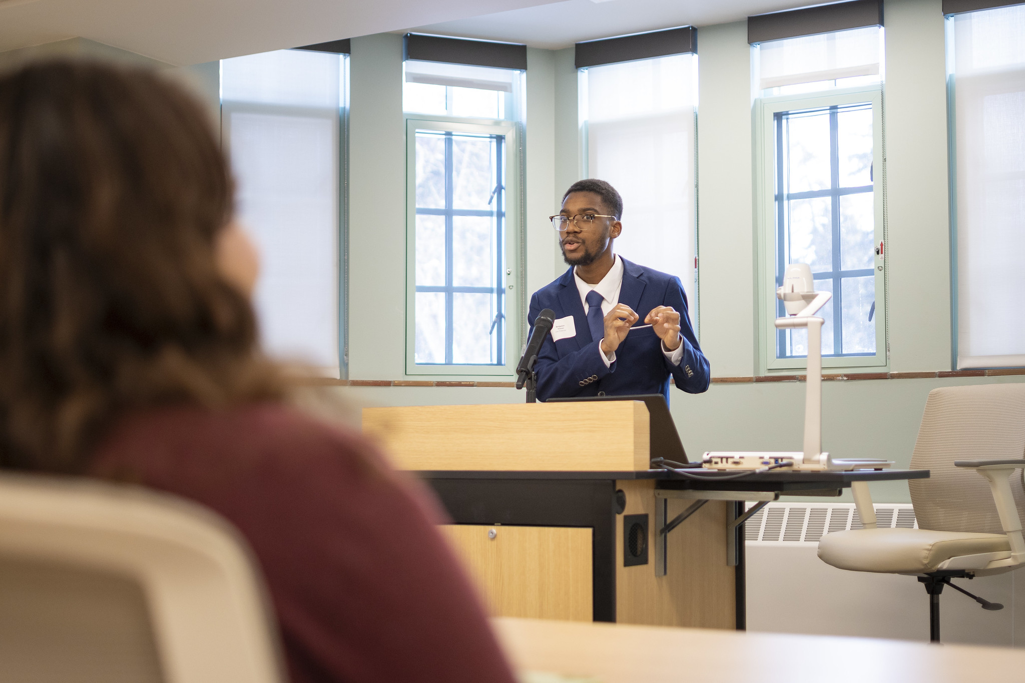 Image of Michigan State University Honors College student Benjamin Nketsiah standing in a classroom and presenting to an audience. Nketsiah is in a navy blue suit and tie, with a white collared shirt.
