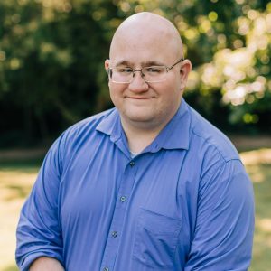 Headshot photo of Bill Chopik in front of greenery, wearing a blue button-up and glasses.