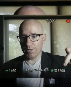 Photo of Troy Hale on a recording camera in the middle of speaking, wearing a white shirt and black sport coat and black-framed glasses.
