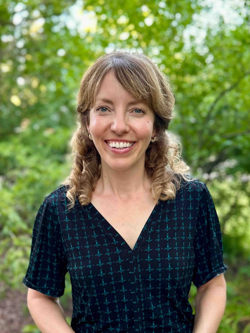 Image of Associate Professor Helen Zoe Veit standing and smiling in a navy and dark green patterned dress. Blurred greenery is in the background.