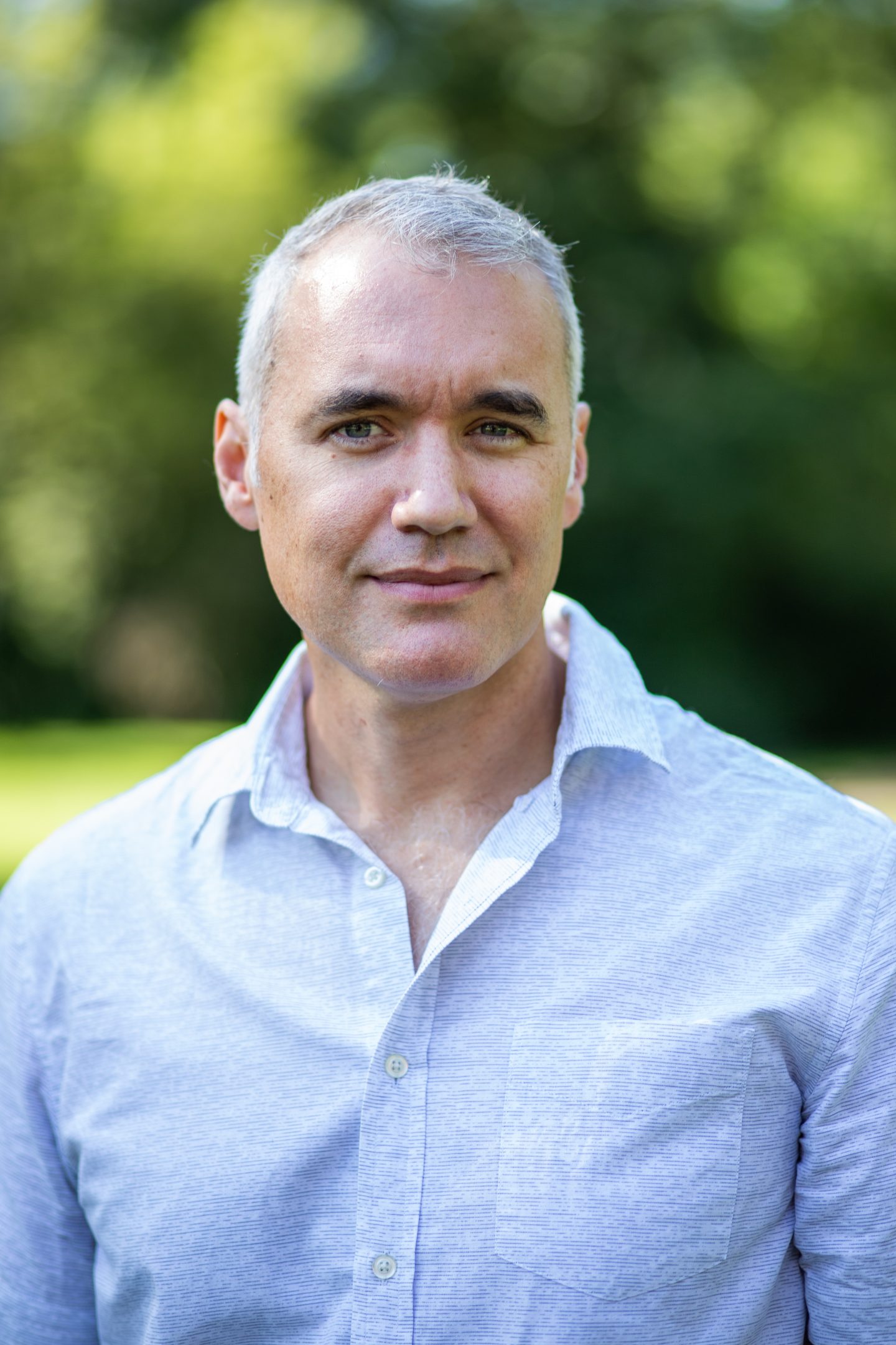 Professional image of Doug Bessette softly smiling in a button-up shirt. Blurred greenery is in the background.