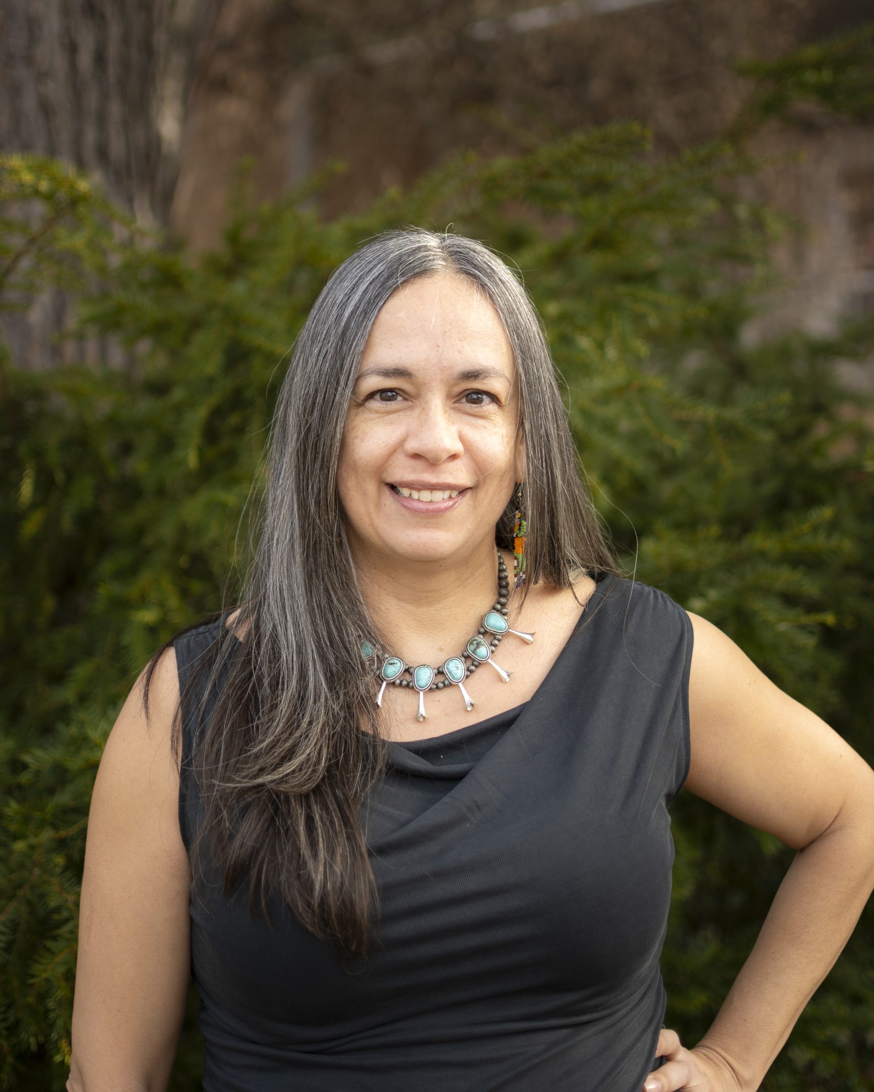 Image of Teresa Dunn standing outside and smiling in front of greenery. Dunn is wearing a black top and beaded necklace with large turquoise stones.