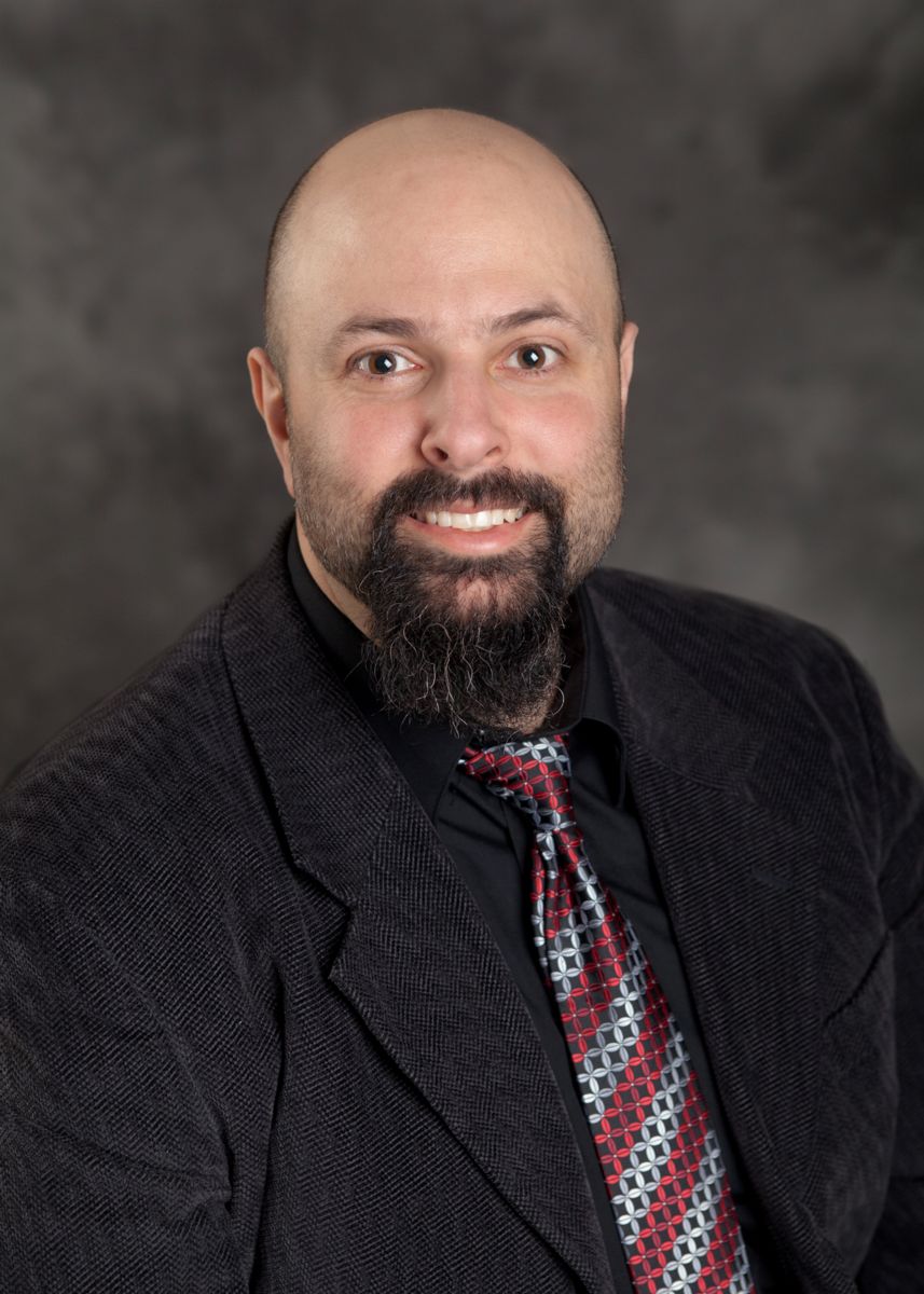 Formal image of Albert Cohen smiling in a black suit, black collared shirt, and silver and red-striped tie against a dark grey gradient background. 