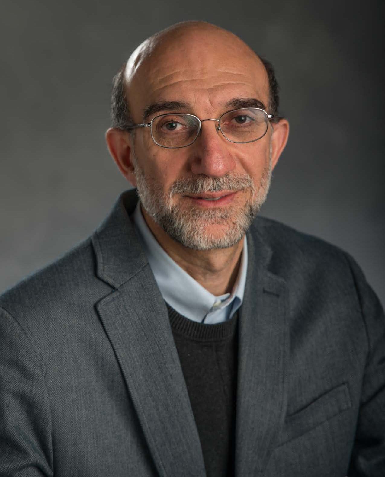 Professional image of Hayder Radha in a grey blazer, dark grey sweater, and collared shirt against a grey gradient background.