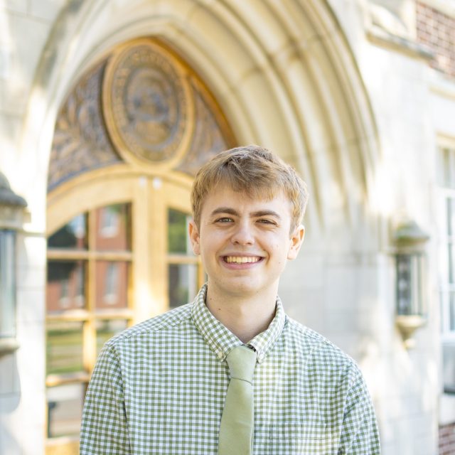 Image of Michigan State University student and debater Glenn Scully standing and smiling in a light green plaid collared shirt and light green tie in front of Linton Hall on MSU’s campus.