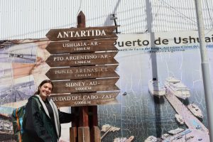 Photo of Julia Preservati standing next to a large wooden sign in a foreign language pointing in different directions for different locations. The biggest text on the sign reads, "Antartida" and points to the left. 