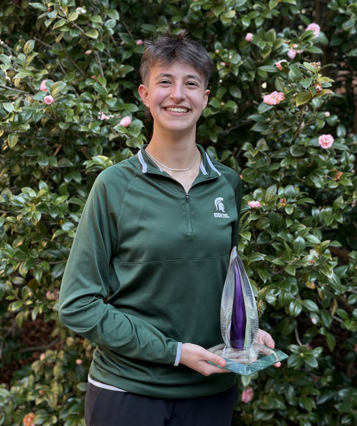 Image of Michigan State University student and debater Joanna Gusis standing and smiling while holding the glass Julia Burke Award. Gusis is wearing a dark green MSU quarter-zip, and flowering greenery is in the background.