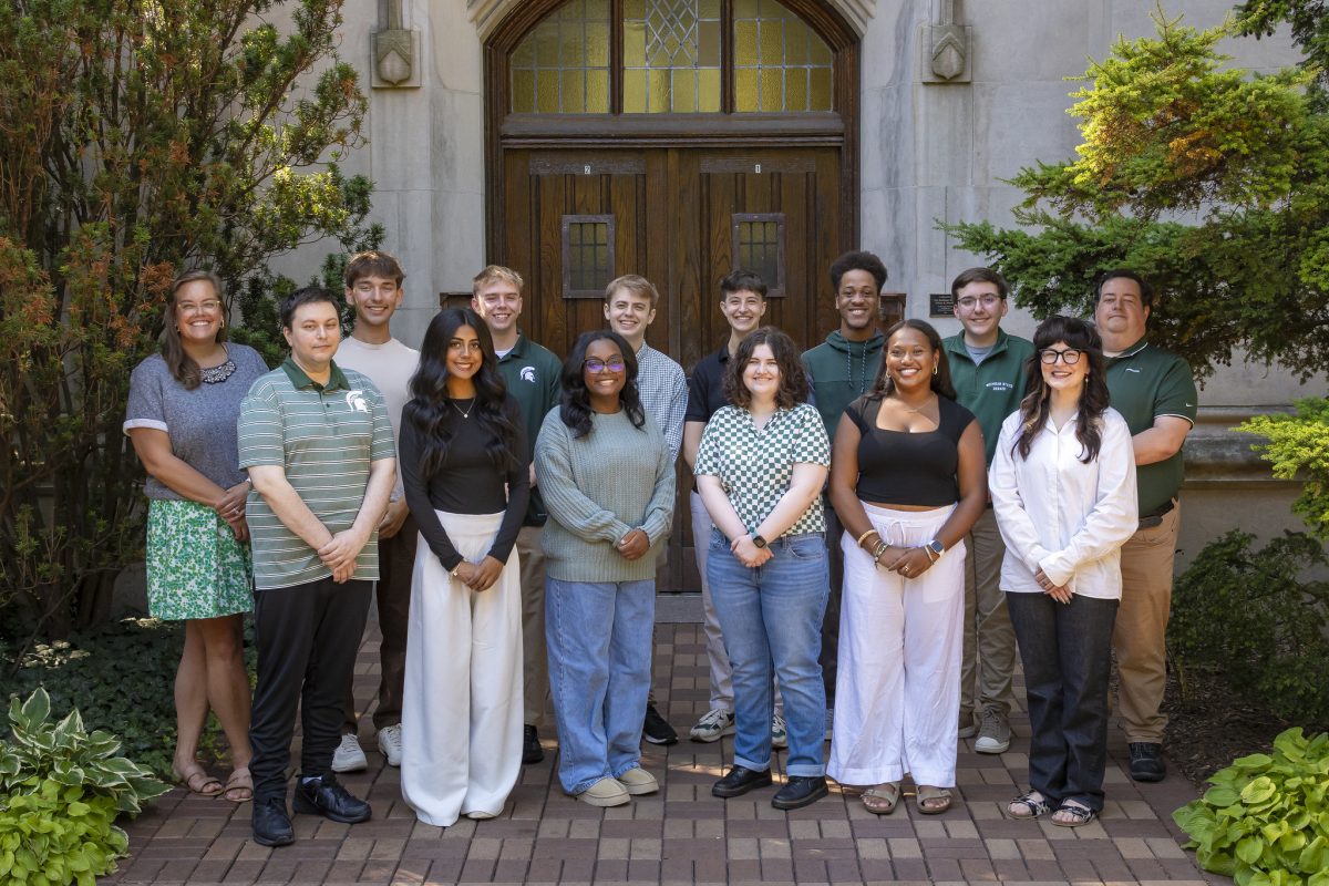 Image of the Michigan State University Debate team and coaches standing in two rows and smiling in front of Beaumont Tower on MSU’s campus. They are wearing various black, white, green, and gray apparel, and are flanked by greenery.