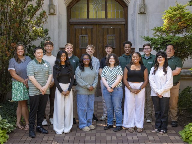 Image of the Michigan State University Debate team and coaches standing in two rows and smiling in front of Beaumont Tower on MSU’s campus. They are wearing various black, white, green, and gray apparel, and are flanked by greenery.