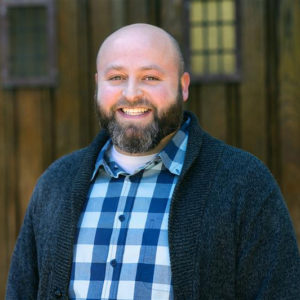 Image of Morgan Shipley standing and smiling in a white and blue plaid collared shirt and dark grey cardigan. Blurred wooden doors are in the background.