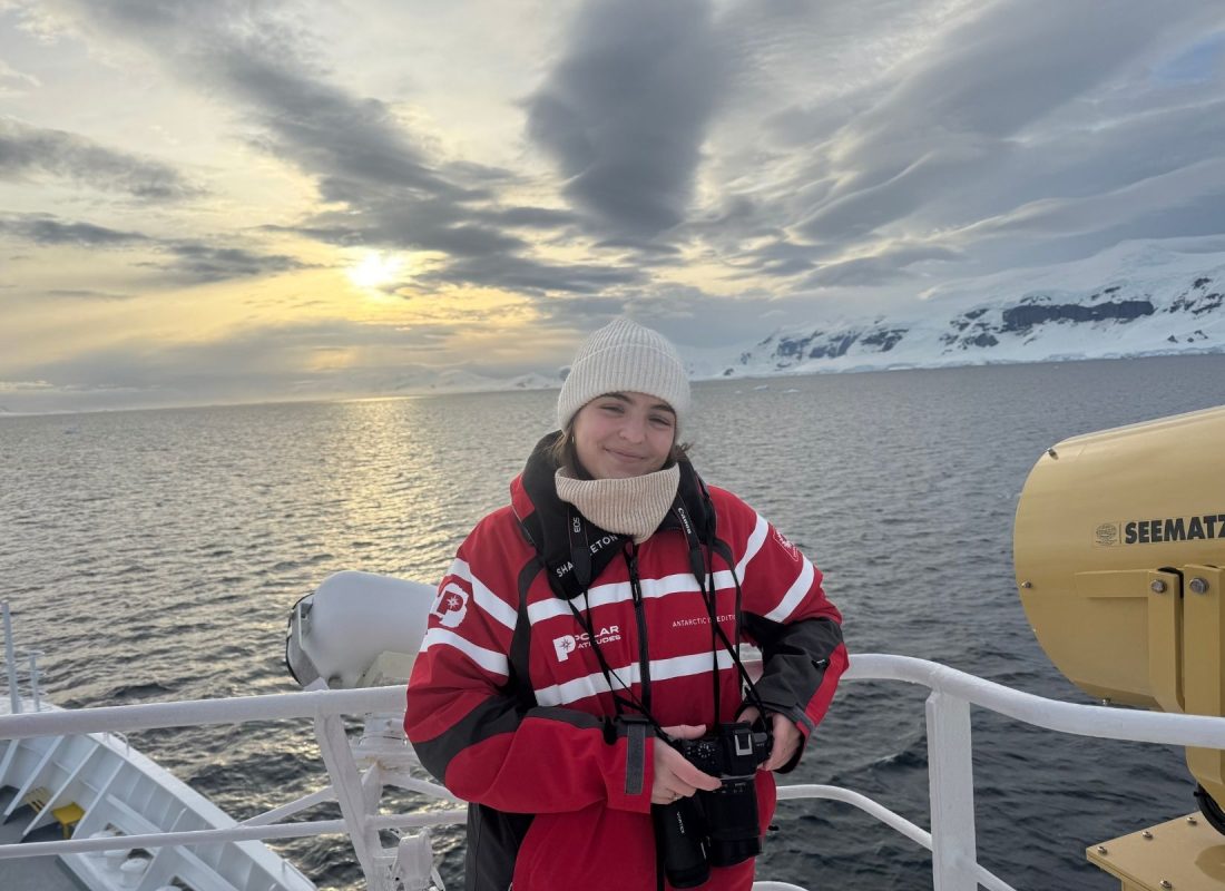 Photo of Julia Preservati wearing a winter coat and holding a camera and smiling on a boat, with a polar ocean in the background.