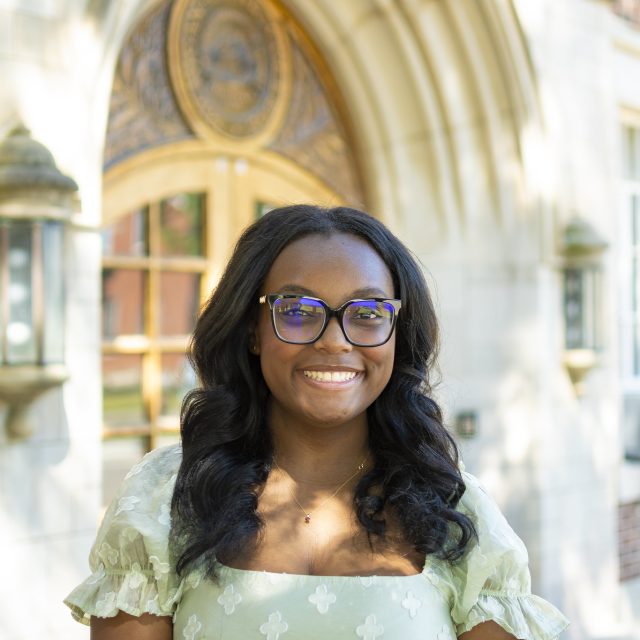 Image of Michigan State University student and debater Zaria Jarman standing and smiling in a light green patterned shirt in front of Linton Hall on MSU’s campus.