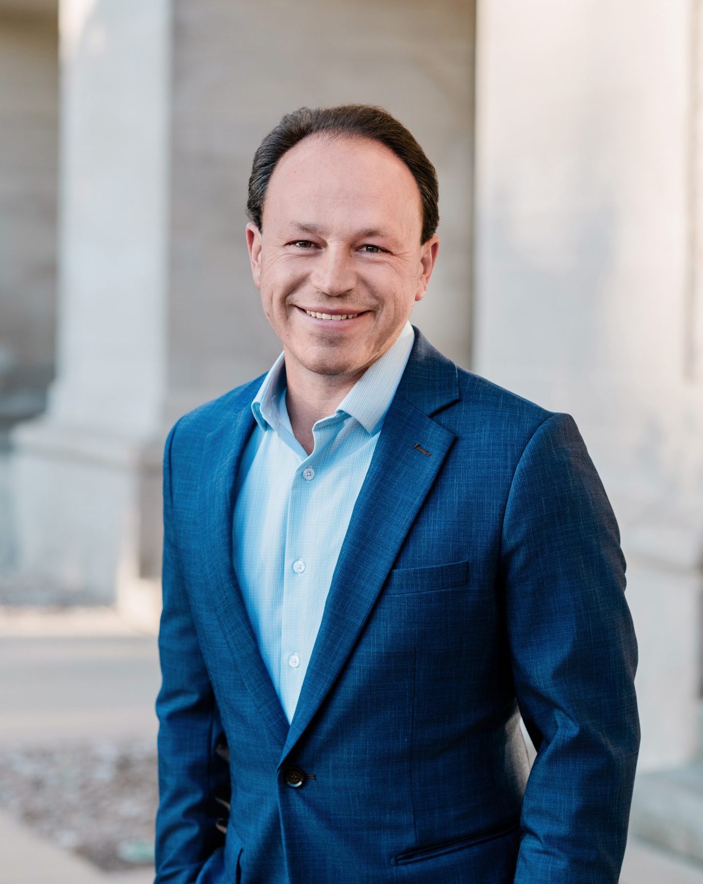 Professional image of Matthew Grossman standing and smiling in a navy blazer and light blue button-up shirt. A blurred columned building is in the background.