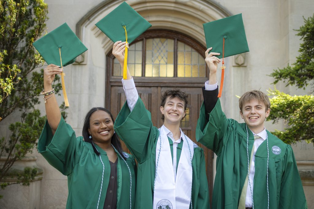 Image of MSU Debate graduates Arielle Gearring, Joanna Gusis, and Glen Scully standing and smiling in their green graduation robes, while holding their green graduation caps to the sky. The wooden door entrance to Beaumont Tower is behind them.