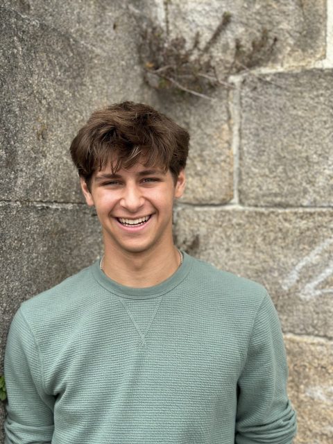 Image of incoming MSU Honors College student and Alumni Distinguished Scholar Gavin Arena, smiling against a stone wall while wearing a sage green long-sleeve shirt.