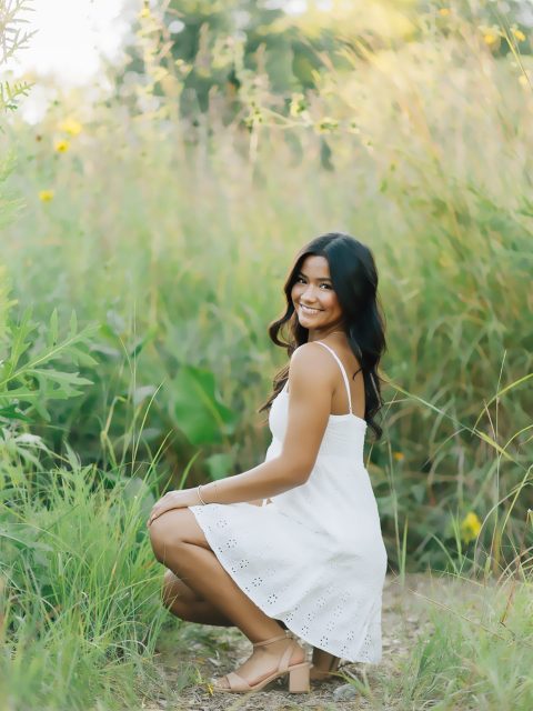 Image of incoming MSU Honors College student and Alumni Distinguished Scholar Miranda Belo smiling in a white sundress and tan heels while crouching down on a grassy trail. Tall grasses and small yellow flowers are in the blurred background.