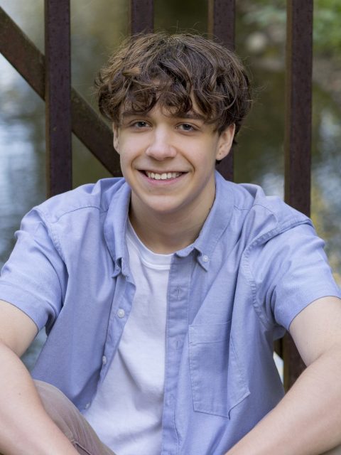 Image of incoming MSU Honors College student and Alumni Distinguished Scholar Giovanni Bickel smiling and sitting against a brown bridge, wearing a short-sleeved blue collared shirt over a white t-shirt. A body of water is in the background.