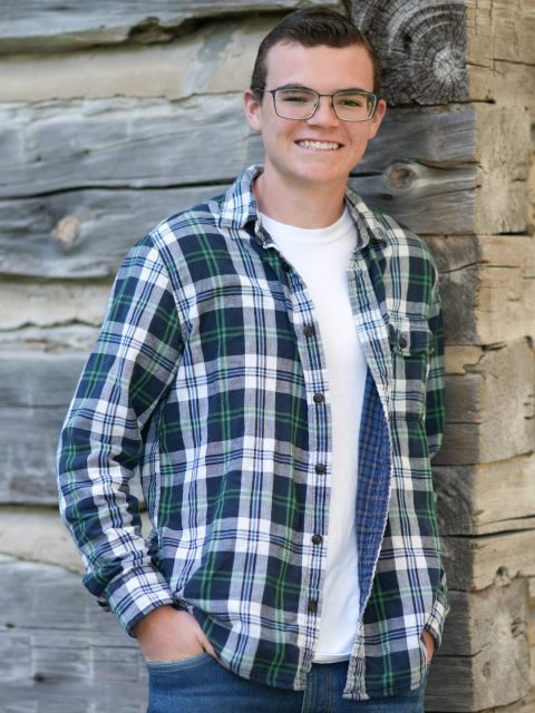 Image of incoming MSU Honors College student and Alumni Distinguished Scholar Carston Broderick standing and smiling against a wooden wall with hands in pockets. Broderick is wearing blue jeans and a green, blue, and white plaid long-sleeve shirt over a white t-shirt.