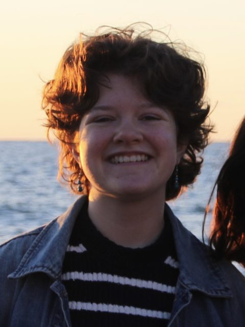 Image of incoming MSU Honors College student and Alumni Distinguished Scholar Fiona Burke smiling in a denim jacket over a striped shirt. A body of water is in the background.