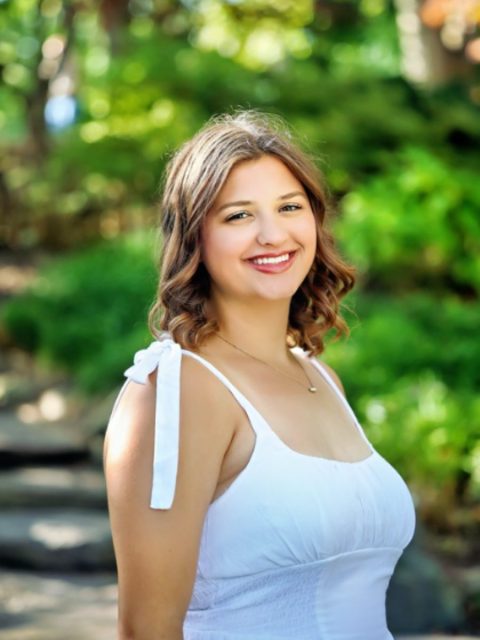 Image of incoming MSU Honors College student and Alumni Distinguished Scholar Avery Collins smiling in a white sundress. Lush greenery is blurred in the background.