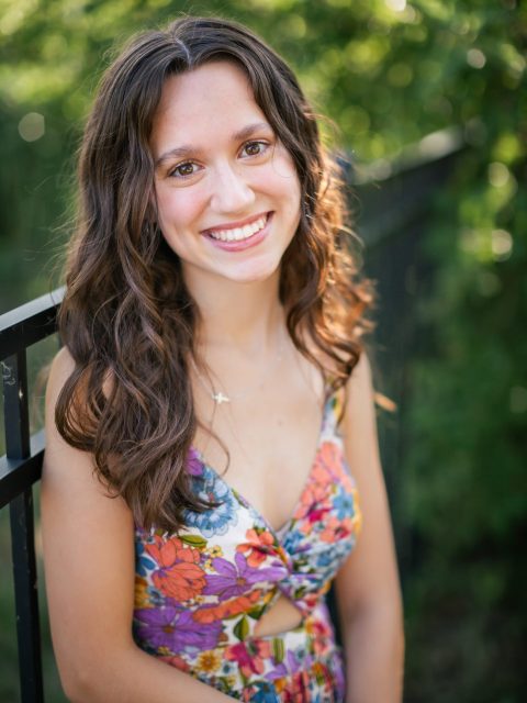 Image of incoming MSU Honors College student and Alumni Distinguished Scholar Kedzie Hall smiling and leaning against a black railing in a colorful floral print dress. Blurred greenery is in the background.