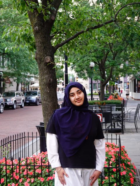 Image of incoming MSU Honors College student and Alumni Distinguished Scholar Maria Khan softly smiling in a navy hijab, wearing a black t-shirt with a white long-sleeve underneath. Trees and flowers line a street of parked cars in the background.