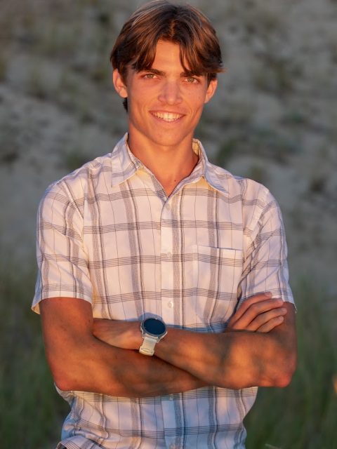 Image of incoming MSU Honors College student and Alumni Distinguished Scholar Lincoln Kuhl standing and smiling with arms crossed. Kuhl is wearing a white watch and white plaid shirt.