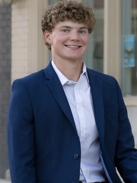 Image of incoming MSU Honors College student and Alumni Distinguished Scholar Evan Maki smiling in a navy suit jacket and white collared shirt. A tan and grey building is in the background.