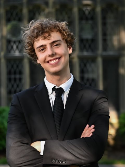 Image of incoming MSU Honors College student and Alumni Distinguished Scholar Peter Moe, smiling with arms crossed. Moe is wearing a black suit jacket, white collared shirt, and black necktie. Blurred greenery and a tan building are in the background.