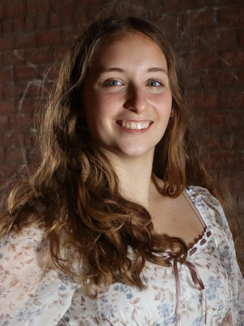 Image of incoming MSU Honors College student and Alumni Distinguished Scholar Rory Welling smiling in a white floral top. A red brick wall is in the background.