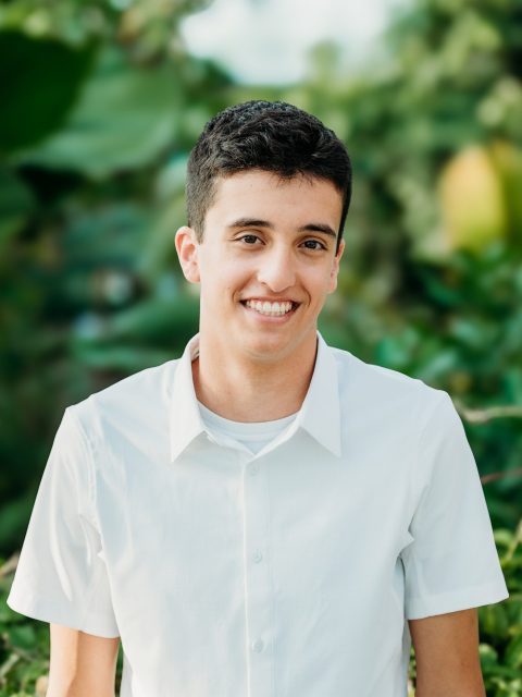 Image of incoming MSU Honors College student and Alumni Distinguished Scholar Eli Zaksek smiling in a white collared shirt. Lush greenery is in the background.