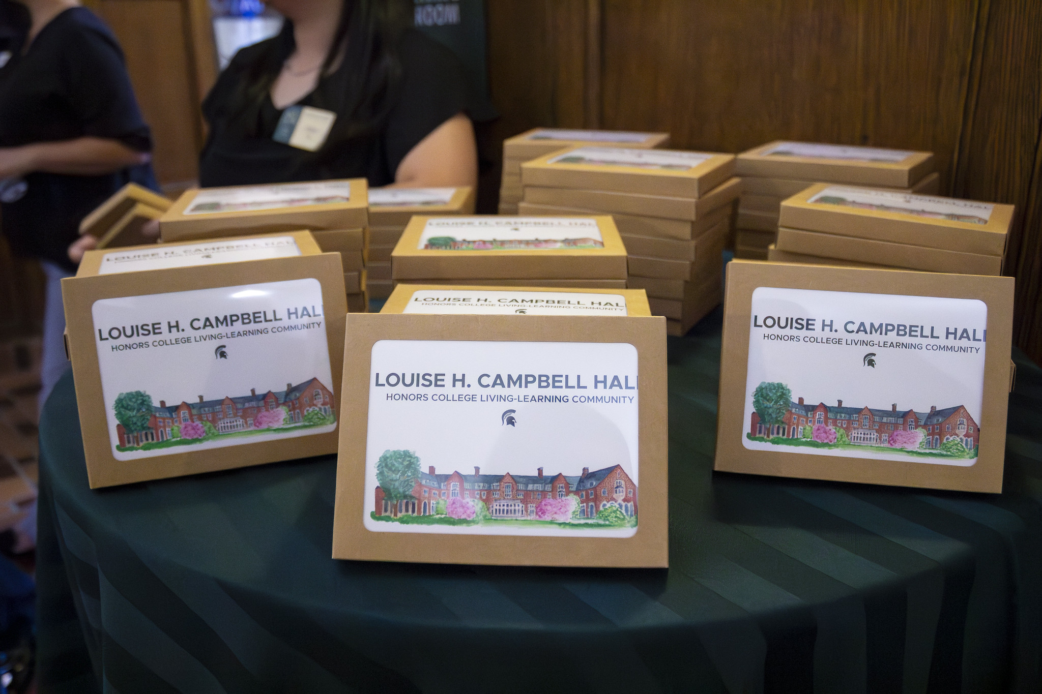 Image of stacks of boxed greeting card sets displayed on a table. Each box features a watercolor of Louise H. Campbell Hall with the text “Louise H. Campbell Hall, Honors College Living-Learning Community.” Two people stand blurred in the background.