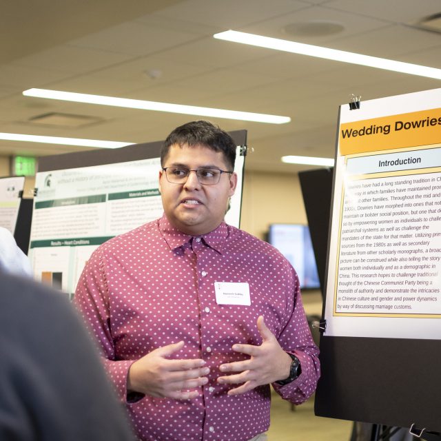 Image of MSU Honors College student Domenic Cedillo presenting a multicolor research poster on wedding dowries. Cedillo is wearing a light red collared shirt and talking. Other posters are in the background.