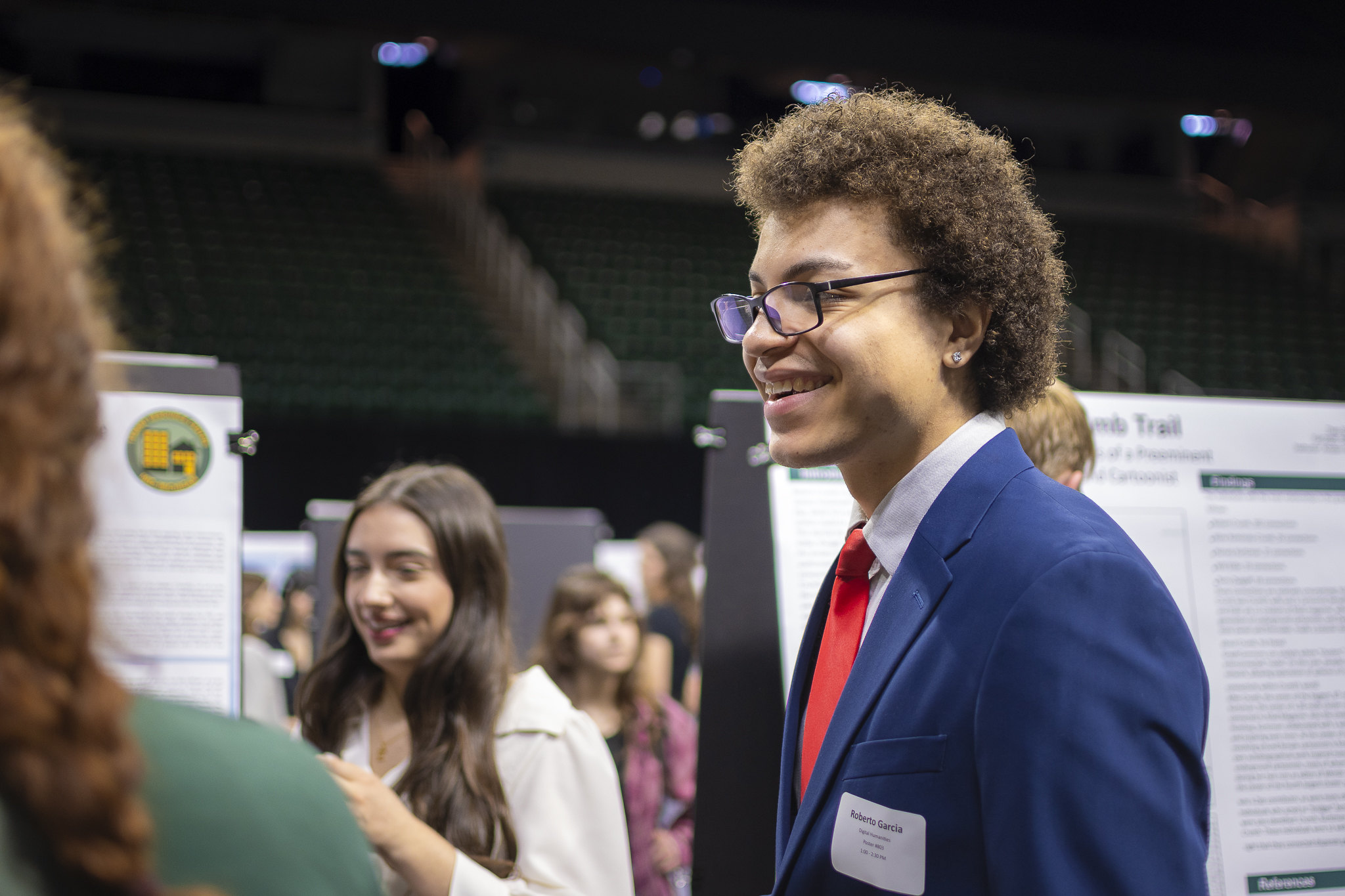 Image of Marcos Garcia standing and smiling in a navy blazer, collared shirt, and red necktie at the Breslin Center on MSU’s campus for the 2026 University Undergraduate Research and Arts Forum. Research posters and other people are in the background.