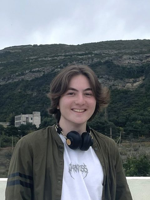 Image of incoming MSU Honors College student and University Distinguished Scholar Andrew Sreseli smiling with headphones around the neck, wearing an olive green jacket and white t-shirt. Tree-covered cliffs are in the background.