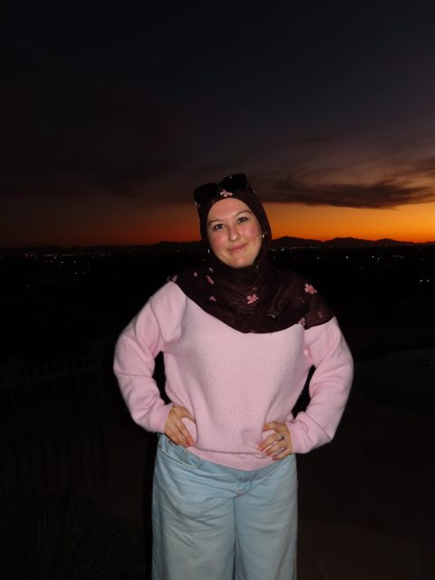 Image of incoming MSU Honors College student and Williams Scholar Safia Behmen standingand softly smiling with hands on hips in a dark floral hijab, light pink sweater, and blue jeans. The afterglow from a sunset is in the background.