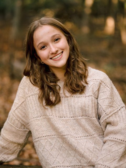 Image of incoming MSU Honors College student and Williams Scholar Katie Foley smiling and leaning toward one shoulder in a wooded area. Foley is wearing an ivory knit sweater.
