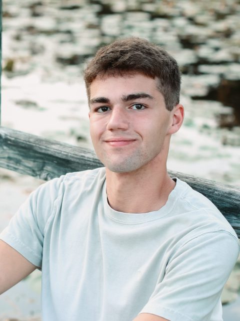 Image of incoming MSU Honors College student and Williams Scholar Ben Holbrook softly smiling and leaning against a wooden railing. Holbrook is in a tan t-shirt, and a body of water is in the background.
