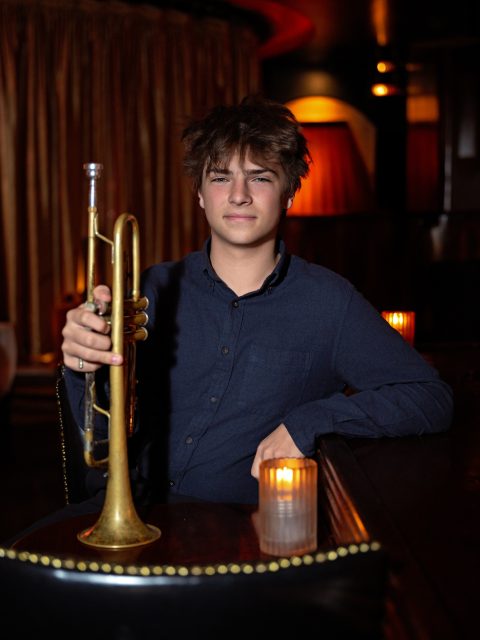 Image of incoming MSU Honors College student and Williams Scholar Casey Knas looking focused while holding a trumpet and wearing a navy collared shirt. A stage is in the background.