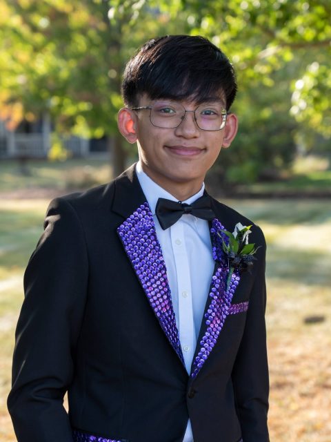 Image of incoming MSU Honors College student and Williams Scholar Kody Leano softly smiling in a tuxedo accented with purple rhinestones. Blurred greenery is in the background.