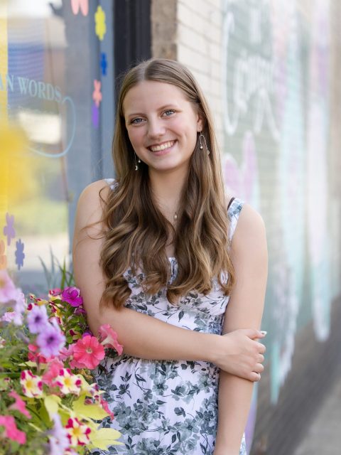 Image of incoming MSU Honors College student and Williams Scholar Greta Lyons standing and smiling in front of flowers and a colorful mural outside a building. Lyons is wearing a green and white floral print dress.